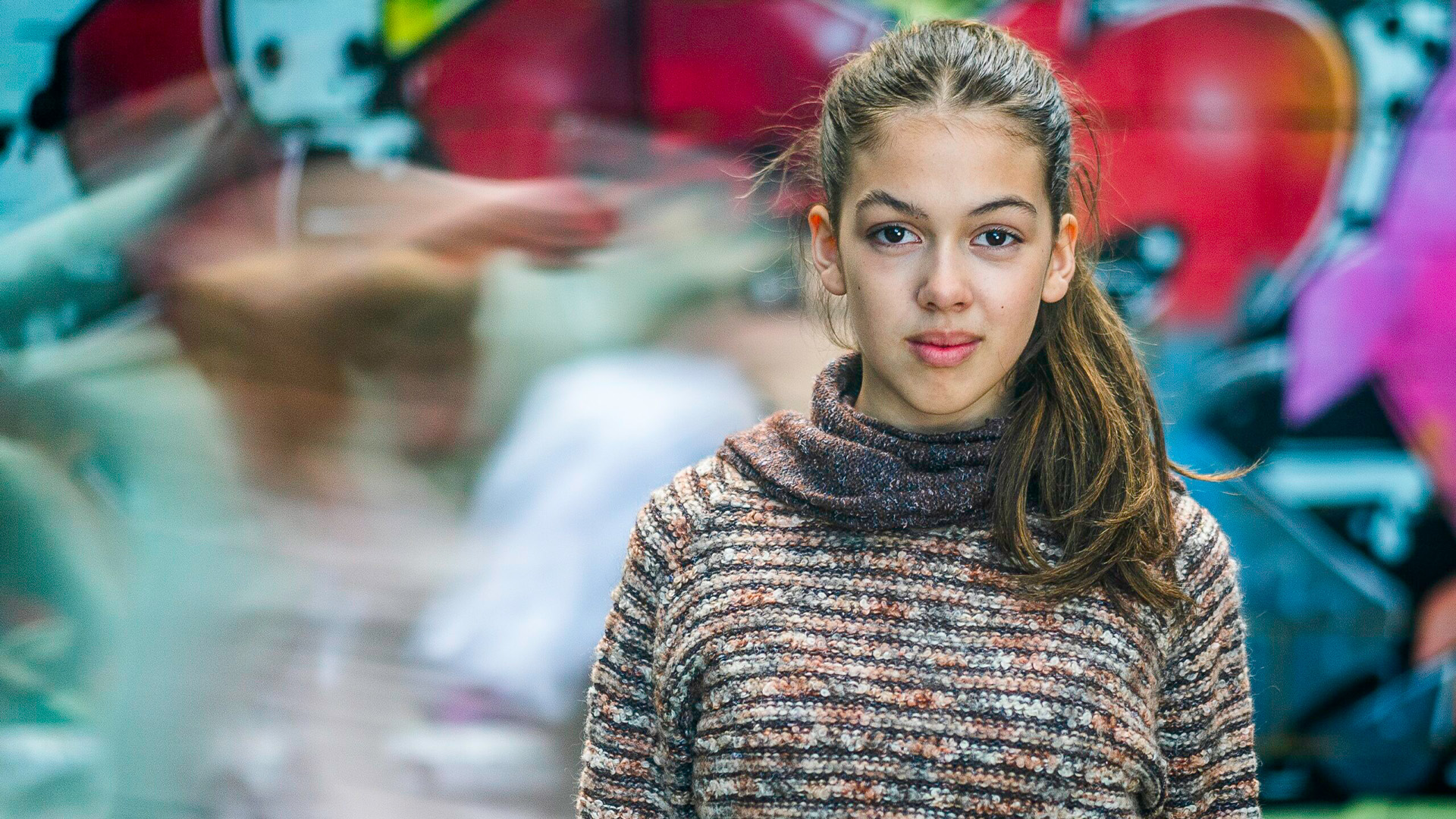 Young woman with a confident expression stands against a colourful graffiti wall. Her hair is in a ponytail, and she wears a striped sweater. Motion blur surrounds her.