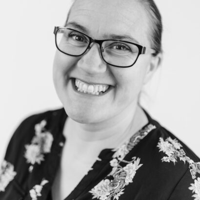 Smiling person wearing glasses and a floral-patterned shirt, captured in black and white. The tone is warm and friendly, with a plain background.