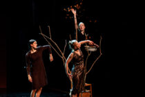 Three performers in dark dresses stand on a dimly lit stage, interacting with bare branches. One raises a hand, releasing leaves, evoking a surreal, artistic atmosphere.