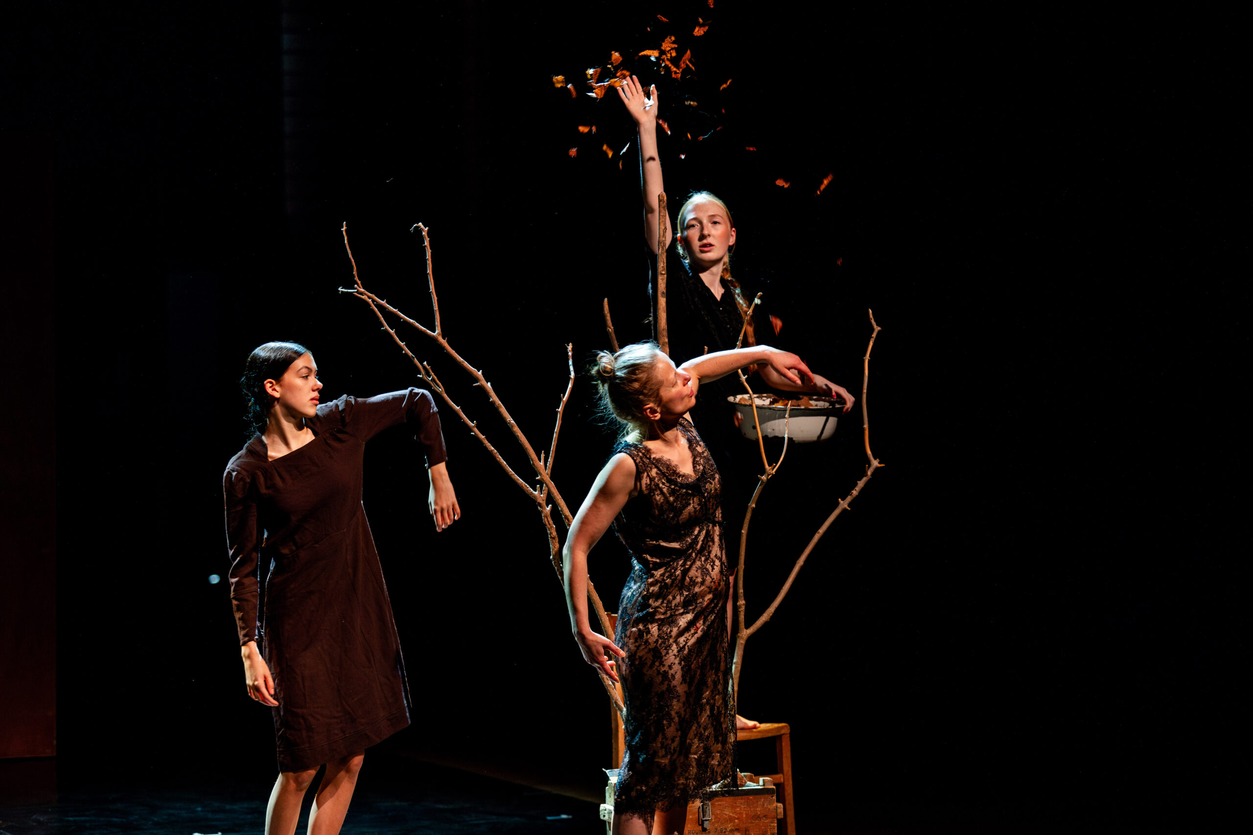 Three performers in dark dresses stand on a dimly lit stage, interacting with bare branches. One raises a hand, releasing leaves, evoking a surreal, artistic atmosphere.