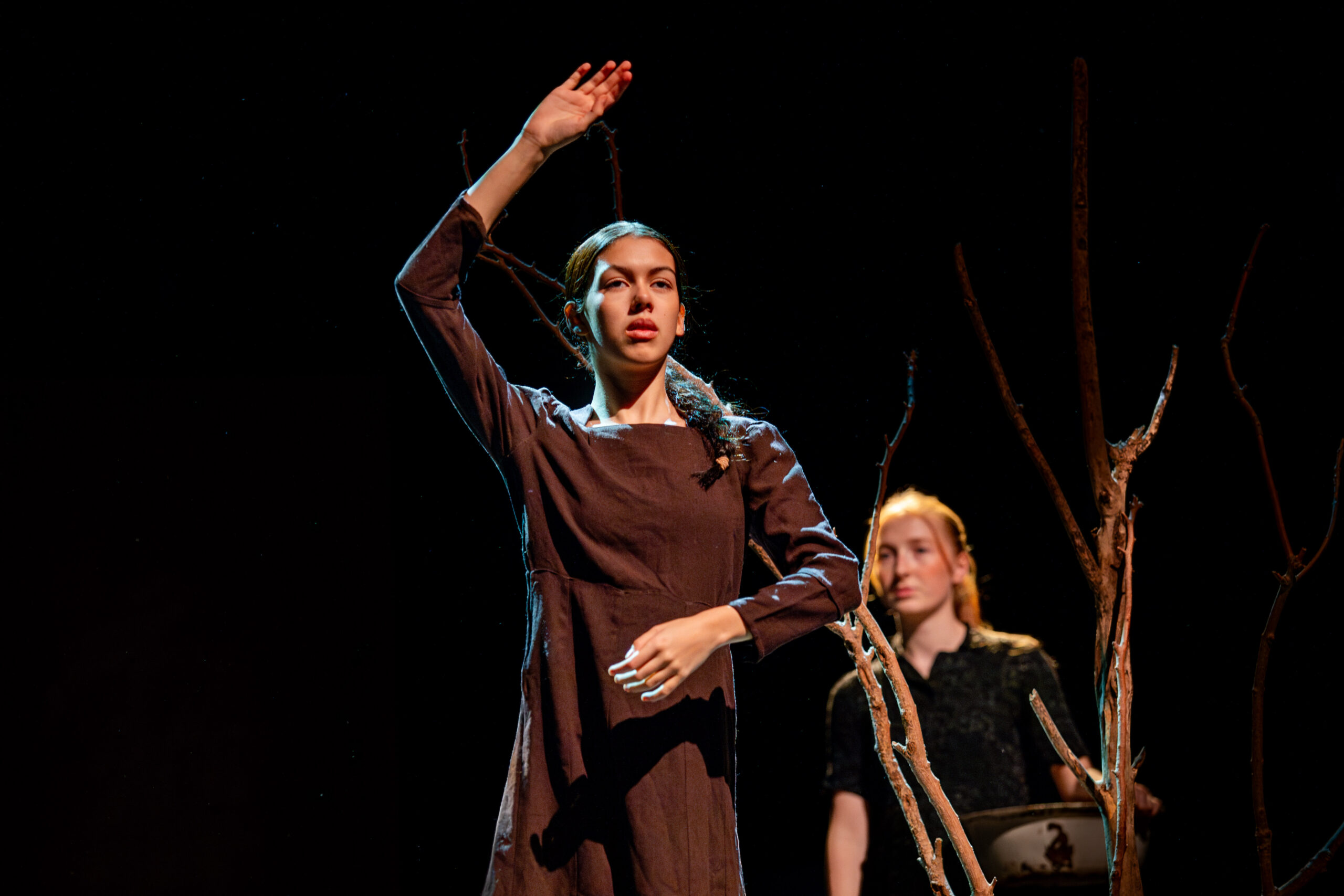 A young woman in a brown dress is on stage, raising one arm, conveying focus and intensity. A second person stands behind, partially obscured by branches.