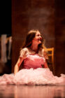 Young girl in a pink dress smiles joyfully while sitting on a stage. A wooden chair is in the background, creating a warm, theatrical ambiance.