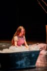 A young girl with long hair, wearing a light pink dress, sits on a dimly lit stage, smiling. A large black tub is in the foreground. The mood is serene and joyful.
