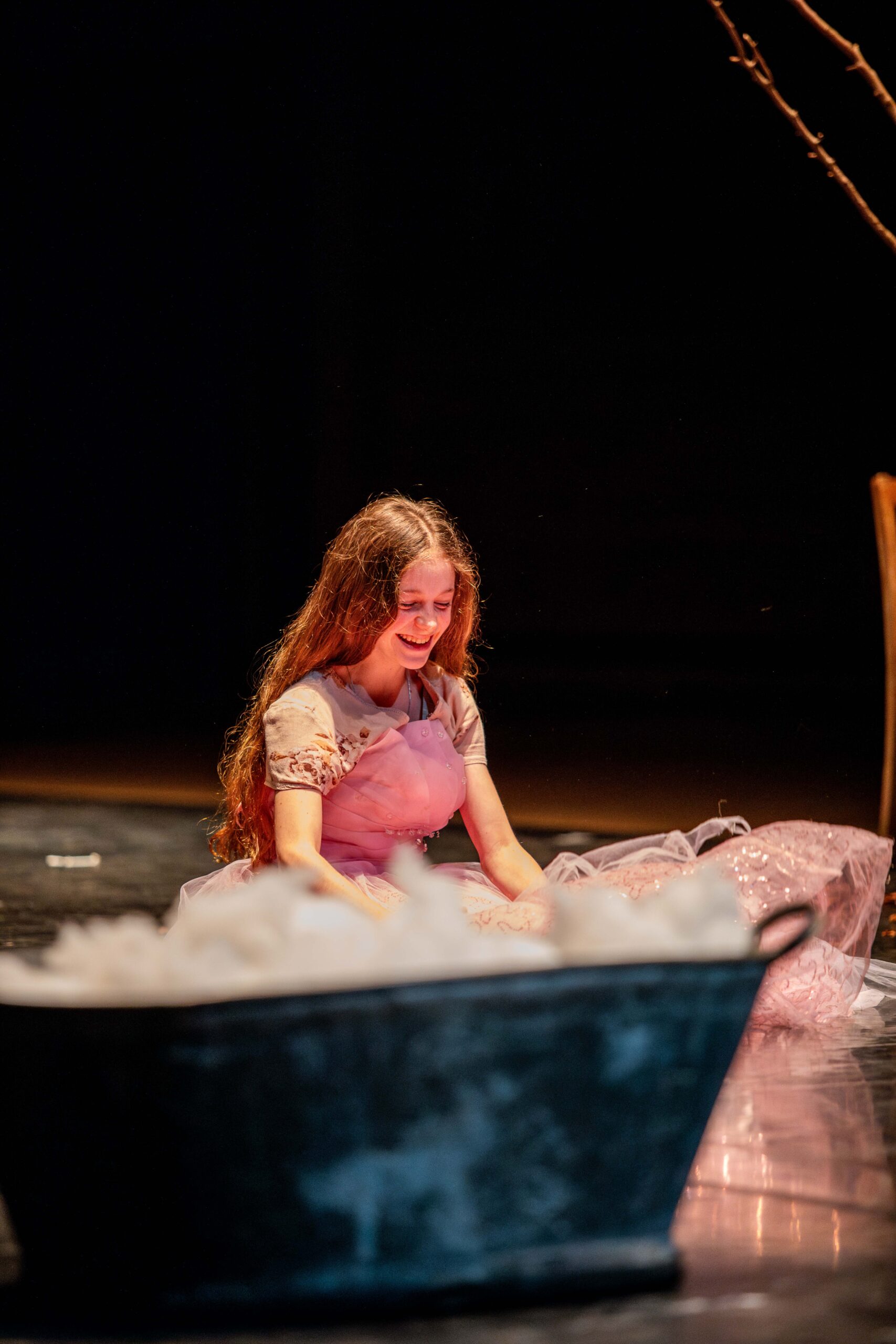 A young girl with long hair, wearing a light pink dress, sits on a dimly lit stage, smiling. A large black tub is in the foreground. The mood is serene and joyful.