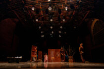 Theatre stage with dramatic lighting, three actors stand amidst minimalist set pieces and bare branches, conveying a tense, moody atmosphere.