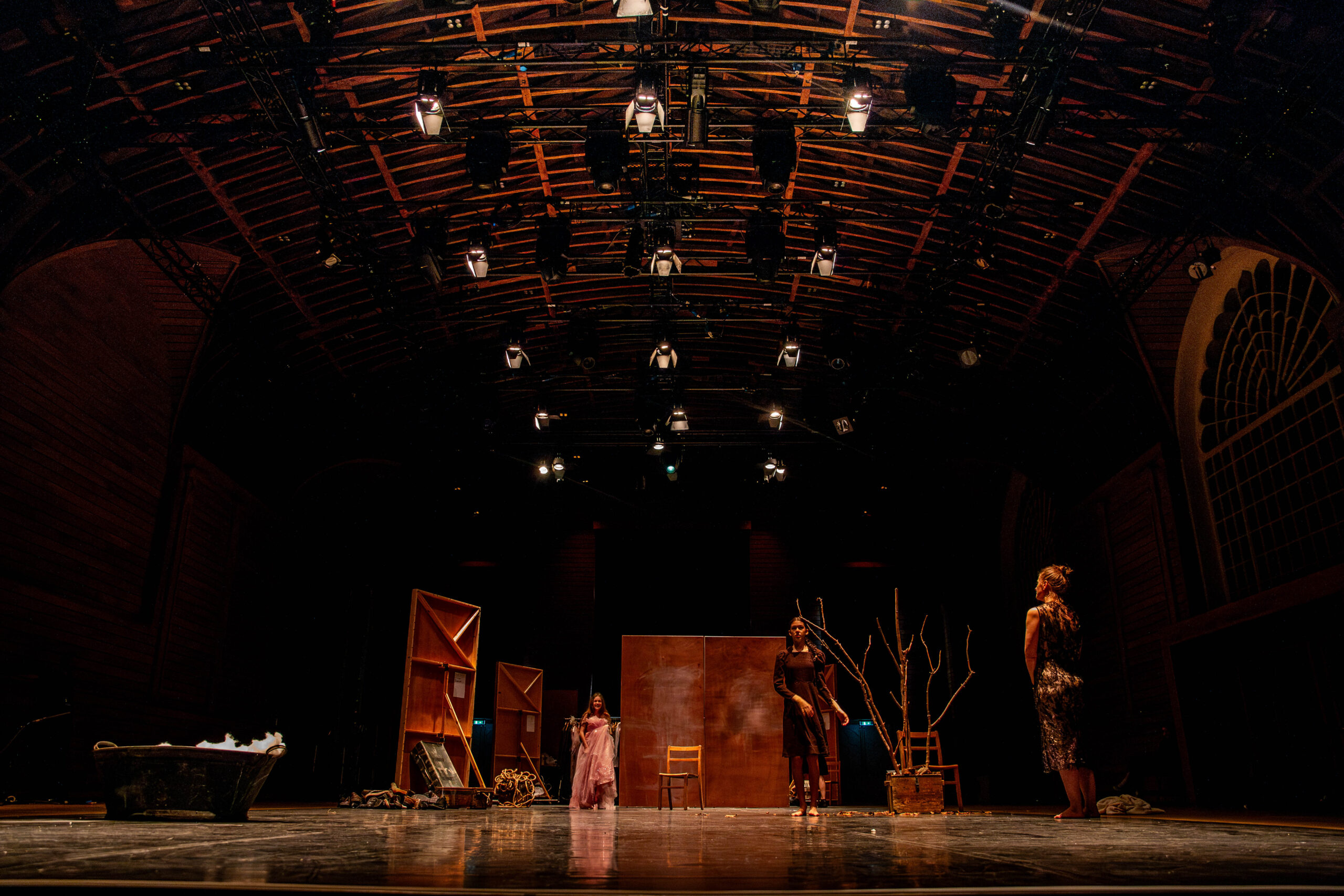 Theatre stage with dramatic lighting, three actors stand amidst minimalist set pieces and bare branches, conveying a tense, moody atmosphere.