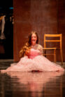 Young girl wearing a pink, sparkly dress sits on a shiny stage floor, smiling joyfully. A wooden chair is behind her, with a dimly lit closet to the side.