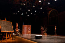 A dimly lit stage featuring two performers standing near bare branches and a large tub. A chalkboard reads, "Let the mountains lead you to love." The atmosphere is contemplative.