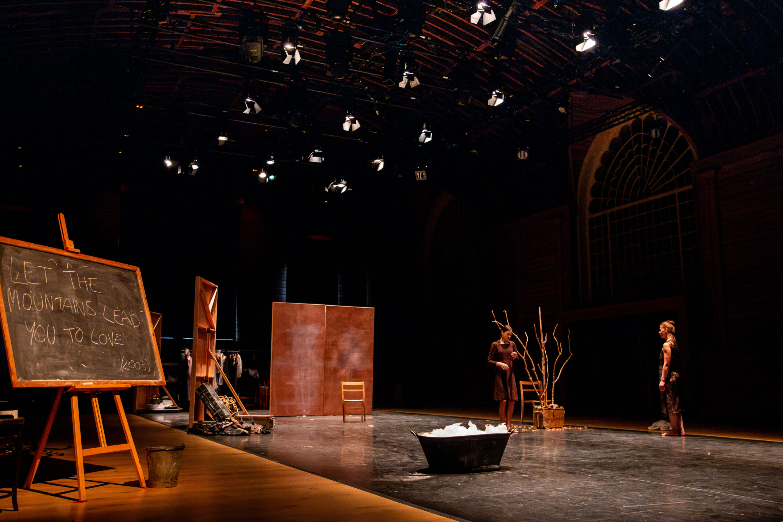 A dimly lit stage featuring two performers standing near bare branches and a large tub. A chalkboard reads, "Let the mountains lead you to love." The atmosphere is contemplative.