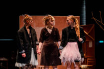 Three women stand on stage, holding hands. They wear mismatched clothing, including black jackets and tutu skirts, conveying a dramatic, tense atmosphere.