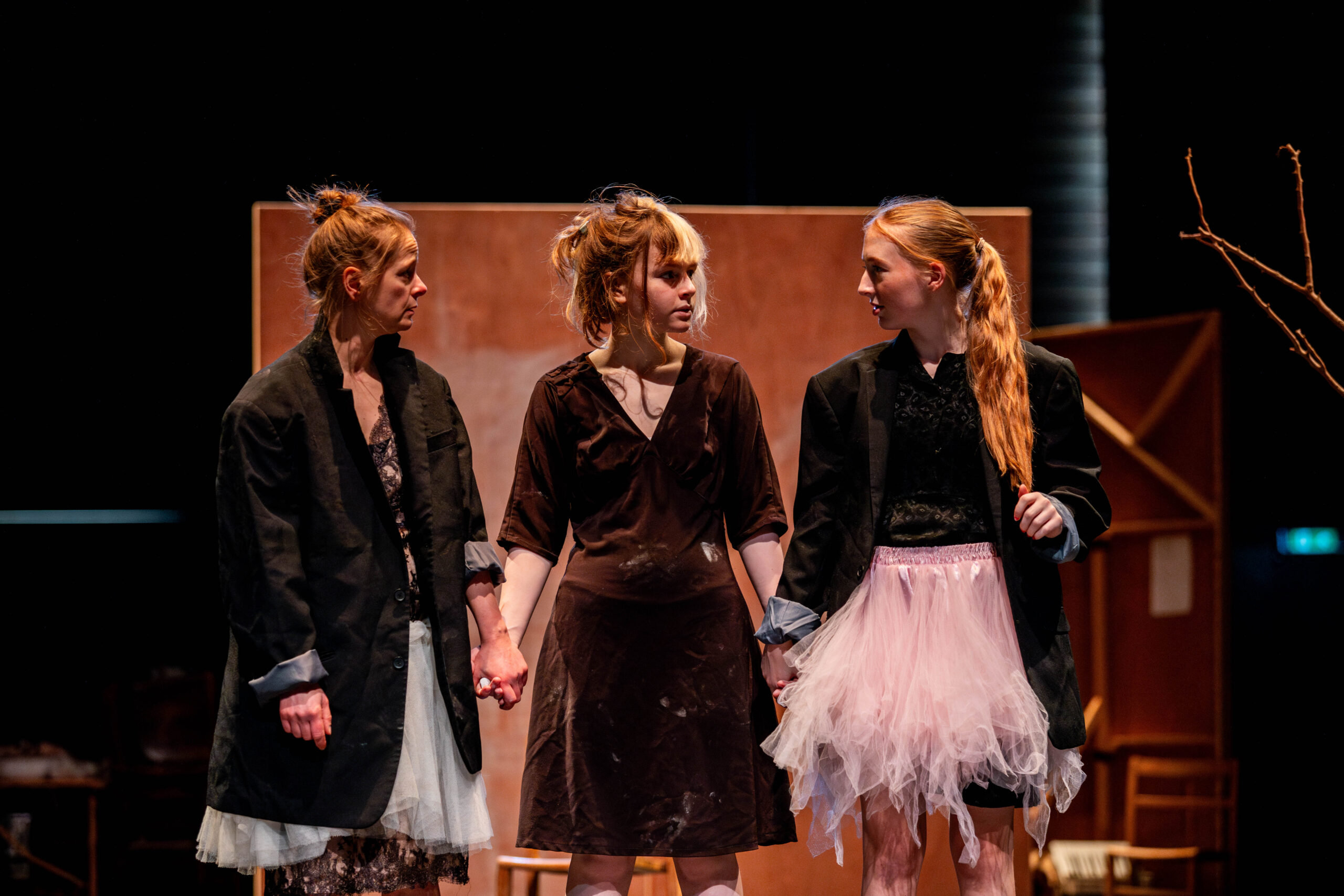 Three women stand on stage, holding hands. They wear mismatched clothing, including black jackets and tutu skirts, conveying a dramatic, tense atmosphere.