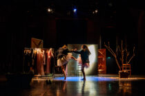 Two dancers in tutus perform under dramatic lighting on a stage. Garment racks and bare branches frame the scene, creating a theatrical, artistic atmosphere.