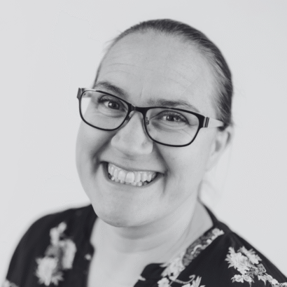 Smiling person wearing glasses and a floral top in a black-and-white portrait; conveys a joyful and friendly tone.