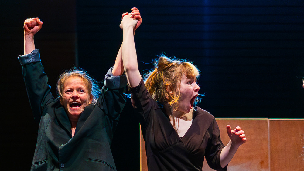 Two women on stage celebrate triumphantly. One raises both fists, smiling, while the other looks shocked and overjoyed as her arm is lifted in victory. The spotlight highlights their emotional expressions.