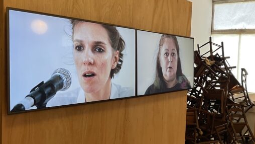 Two large screens display close-up images of people speaking into a microphone, with a backdrop of stacked wooden chairs.