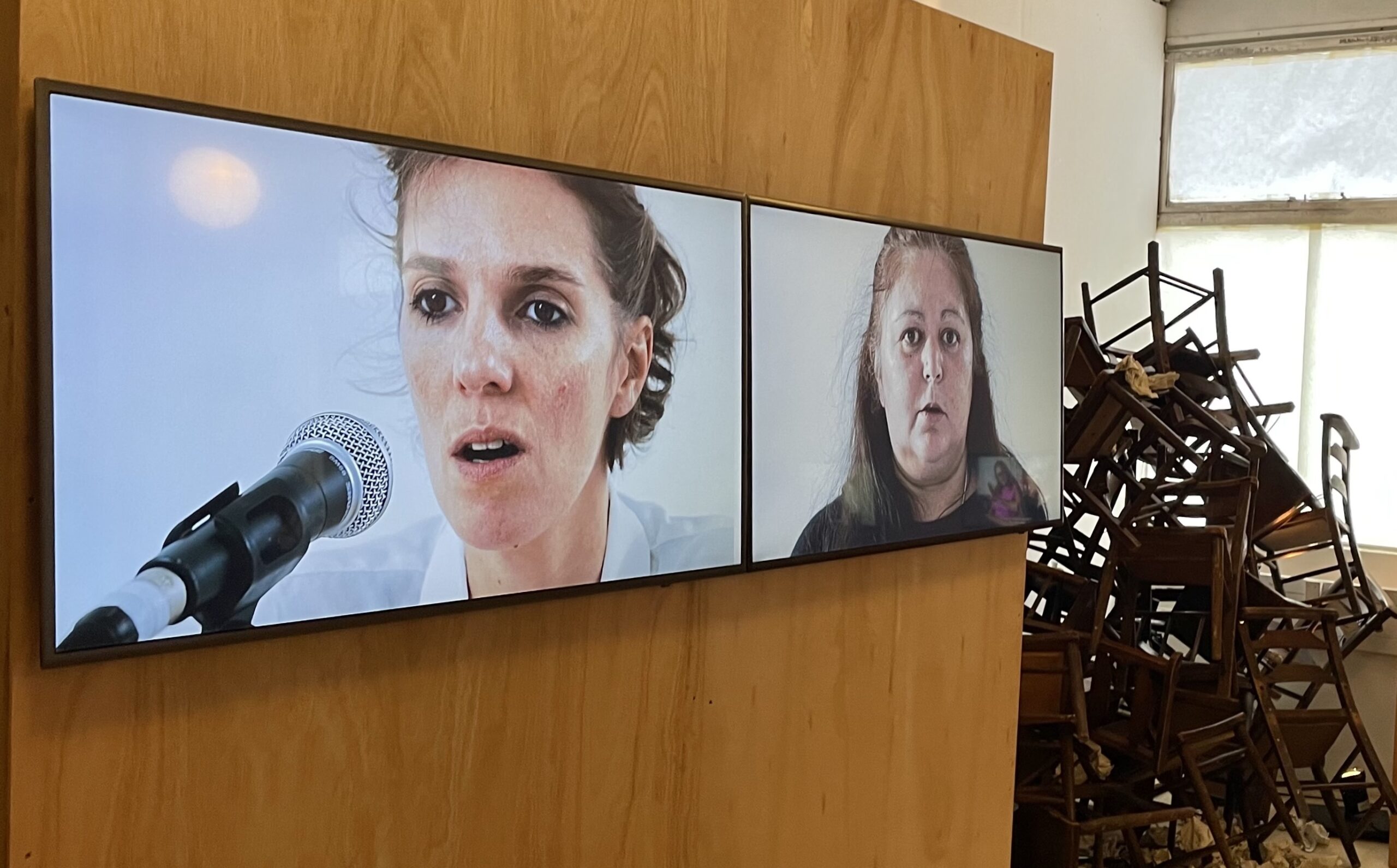 Two large screens display close-up images of people speaking into a microphone, with a backdrop of stacked wooden chairs.