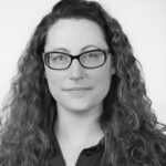 A woman with long, curly hair is wearing a dark button-up shirt, set against a plain white background in a monochrome style.