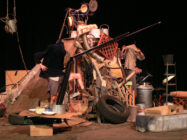 A cluttered stage set with piles of junk, including tires, metal scraps, and a bicycle. Two performers interact creatively, evoking a chaotic yet whimsical atmosphere.