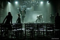 Dimly lit room with six people dynamically moving between rows of stacked chairs. Dramatic lighting and fog create an intense, theatrical atmosphere.