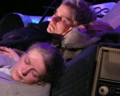 Two people are sleeping against one another, surrounded by various objects in a dim setting. Their expressions appear peaceful and tired. A vintage radio is visible in the foreground.