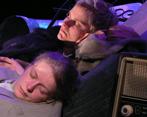 Two people are sleeping against one another, surrounded by various objects in a dim setting. Their expressions appear peaceful and tired. A vintage radio is visible in the foreground.