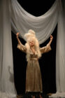 A person in an elaborate white wig and gold robe stands with raised arms on a stage, framed by billowing white curtains, creating a dramatic, theatrical scene.