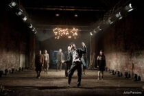 A dramatic stage scene shows a man in a suit dancing exuberantly under a chandelier in a dimly lit, rustic setting, with five people watching behind him.