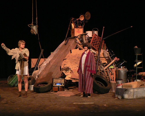 Two actors stand on stage amidst a chaotic pile of junk, evoking a sense of absurdity. One wears casual clothes, the other a long coat and hat.