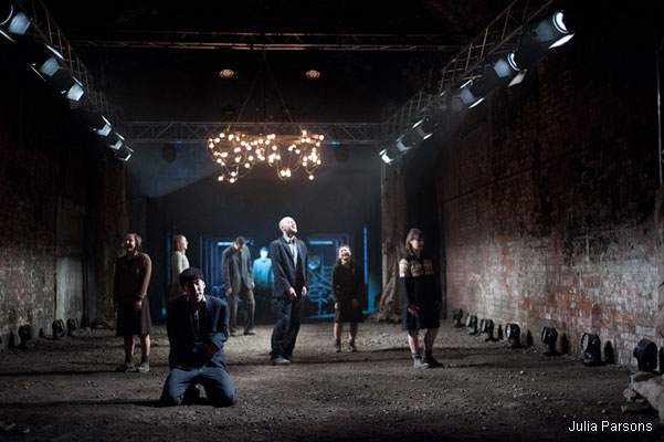 A dimly lit scene shows seven people in an industrial setting. A man kneels in the foreground. The mood is tense and dramatic, with exposed brick walls and scaffolding.