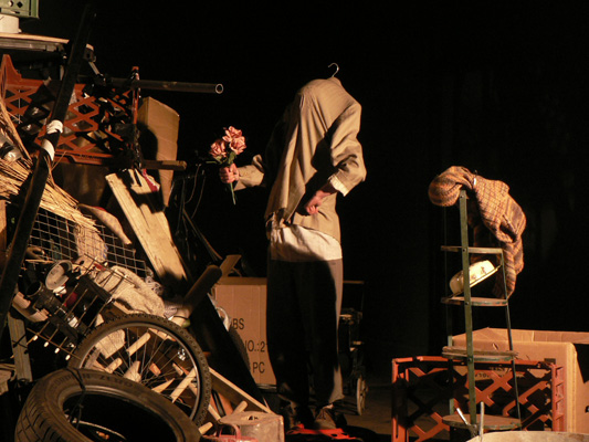A headless mannequin with a shirt holds flowers in a cluttered space with tires, crates, and boxes. Dim lighting creates an eerie, surreal atmosphere.