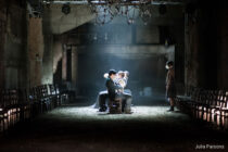 Dimly lit, industrial theater scene with three actors seated center-stage, wearing masks. Empty chairs line the sides. A sombre, atmospheric mood.