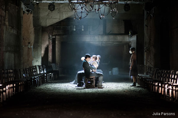 Dimly lit, industrial theater scene with three actors seated center-stage, wearing masks. Empty chairs line the sides. A sombre, atmospheric mood.