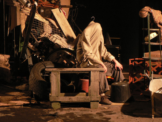 A figure with a coat over their head sits on a wooden bench among scattered items, including tires, crates, and pots, creating a sombre, chaotic scene.