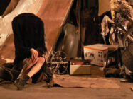 A person sits hunched in a dark coat, head hidden, in a cluttered space with scattered objects, including a vintage radio and a bicycle wheel, conveying isolation.