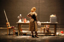 A woman in a patterned dress stands at a cluttered table on a dimly lit stage. The table is strewn with kitchen items and debris, conveying chaos and contemplation.
