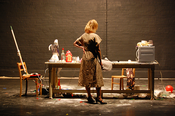 A woman in a patterned dress stands at a cluttered table on a dimly lit stage. The table is strewn with kitchen items and debris, conveying chaos and contemplation.