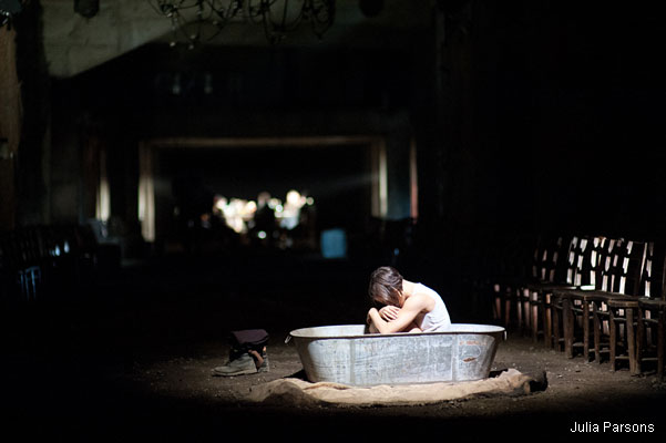 A lone person sits curled up in an empty metal tub under a spotlight in a dimly lit room, surrounded by chairs, evoking feelings of solitude and melancholy.