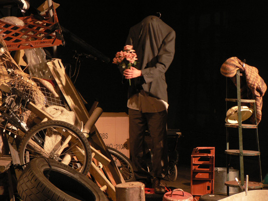 A headless figure in a suit holds pink flowers amid a cluttered scene of stacked tires, wooden items, and ladders, creating an eerie, surreal atmosphere.