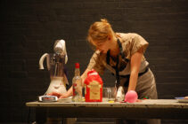 A woman in a beige dress leans over a kitchen table with baking ingredients, including flour and a mixer. The setting suggests a focused, creative atmosphere.