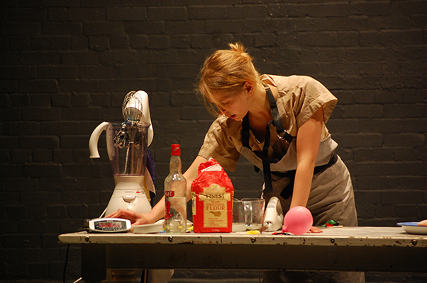 A woman in a beige dress leans over a kitchen table with baking ingredients, including flour and a mixer. The setting suggests a focused, creative atmosphere.