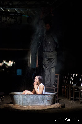 A person sits anxiously in a metal tub while water pours down from above, surrounded by dim light and empty chairs, creating a tense, dramatic atmosphere.