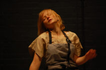 A person in a beige top and gray apron moves expressively against a dark backdrop, eyes closed, conveying a sense of emotion or performance.