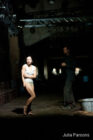 A dimly lit stage shows a woman in minimal clothing, appearing tense or cold, standing near a man in dark clothing. The scene conveys a sombre, dramatic tone.
