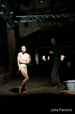 A dimly lit stage shows a woman in minimal clothing, appearing tense or cold, standing near a man in dark clothing. The scene conveys a sombre, dramatic tone.
