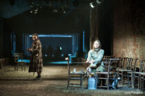 Two women are in a dimly lit, rustic theater set. One sits winding yarn on a spinning wheel, while the other stands, looking contemplative. The scene evokes a sombre, introspective mood.