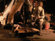 A headless figure in a suit holds pink flowers amid a cluttered scene of stacked tires, wooden items, and ladders, creating an eerie, surreal atmosphere.