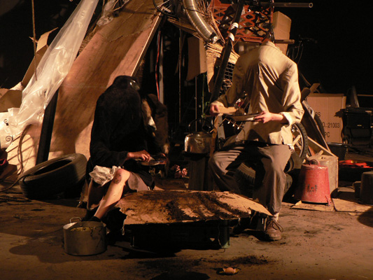 A headless figure in a suit holds pink flowers amid a cluttered scene of stacked tires, wooden items, and ladders, creating an eerie, surreal atmosphere.