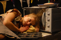A woman rests her head on a cloth beside a microwave, looking contemplative. Nearby are muffin tins, flags, and a plate of food, suggesting a kitchen setting.