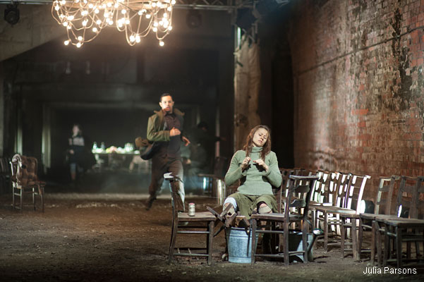 A woman in a green dress knits while sitting on a chair in a dim, rustic room with brick walls. A man runs past under a chandelier, adding tension.