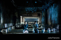 Two individuals kneel in a dimly lit theater with chairs lining the sides. The atmosphere is sombre, with cool blue lighting and shadows.